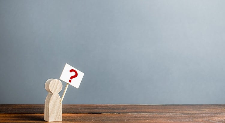 A small wooden figure with a question mark sign, placed on a wooden surface against a plain background. This image represents questioning or decision-making.