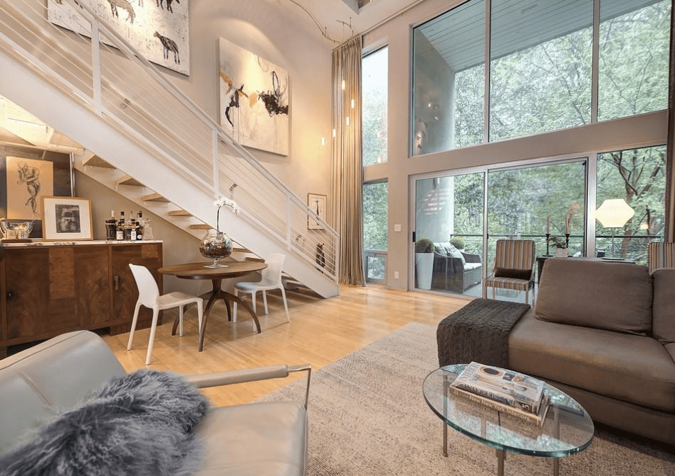 A living room with a glass coffee table, plush couch, metal staircase, light wood treads, white walls, and gray carpet.