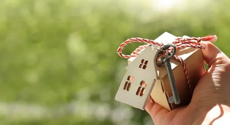 A hand holding a small house keychain with a miniature house attached to it. The background is blurred greenery, suggesting a real estate or homeownership theme.