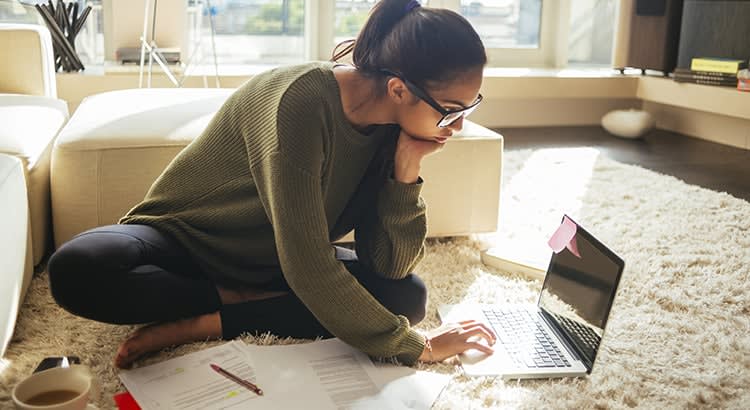 A woman sits on the floor, deeply focused on her laptop, surrounded by papers and books. This suggests a serious work or study session, perhaps a home office or academic setting.