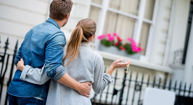 A couple standing outside of what appears to be a newly purchased home. They are shown from behind, with the man’s arm around the woman as they look at the house. 