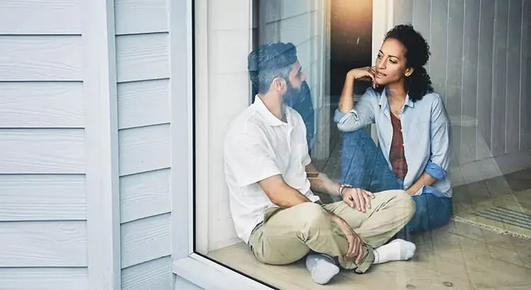 A man and a woman sitting on the floor inside a house, near a large window. 