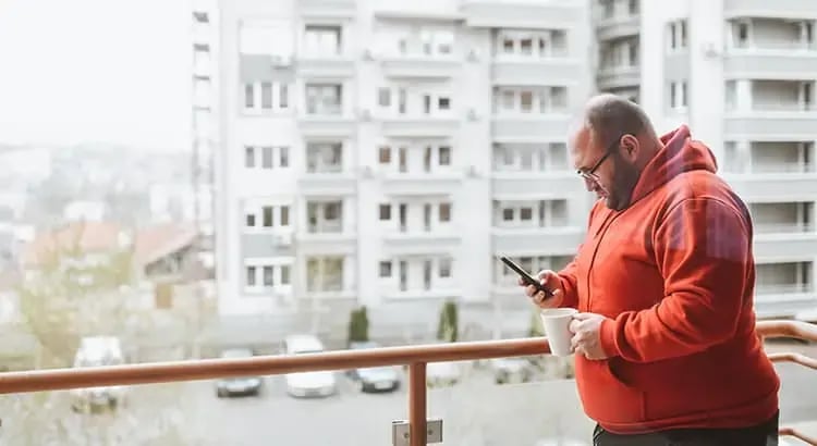 A man in a red hoodie standing on a balcony, looking at his phone. The background shows a cityscape with tall buildings, suggesting an urban setting.