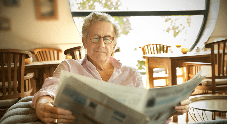 An elderly man comfortably seated indoors, reading a newspaper.