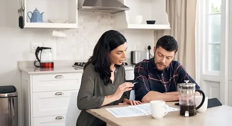 A couple sitting at a kitchen table, reviewing documents together. They appear to be discussing finances or planning something important. The setting is a modern kitchen with white cabinetry.