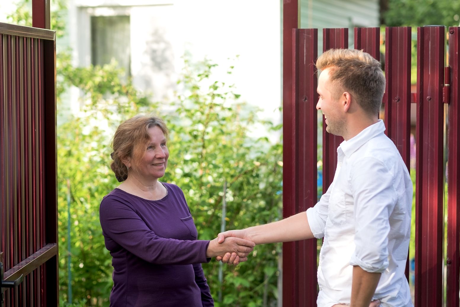 Neighbors smile and shake hands at an open red gate surrounded by greenery.