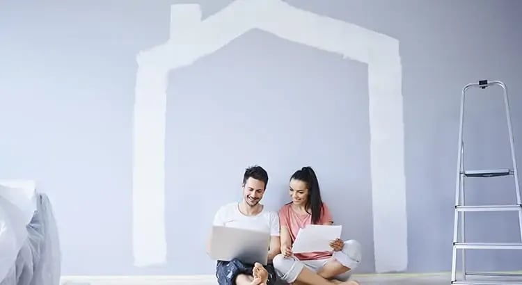 A couple sitting on the floor of a room, holding a house-shaped cutout. There are painting supplies and a ladder nearby, suggesting they are renovating or decorating their home.