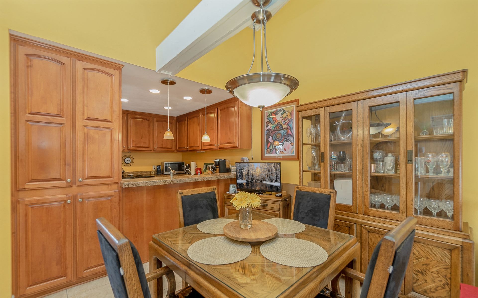 Dining area with a glass-top table, wood chairs, display cabinet, and adjoining kitchen.