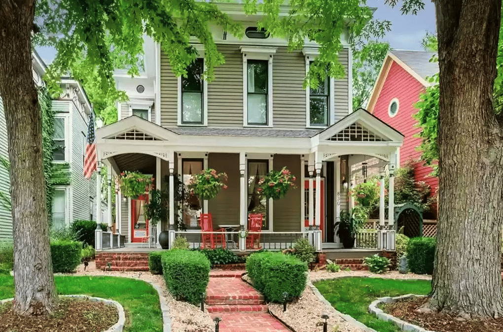 Two-story gray house with a covered porch, red chairs, hanging flowers, and a brick front walkway.