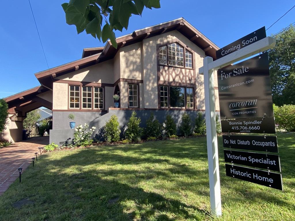 A charming Victorian house with a for sale sign in front surrounded with trees and lawn.