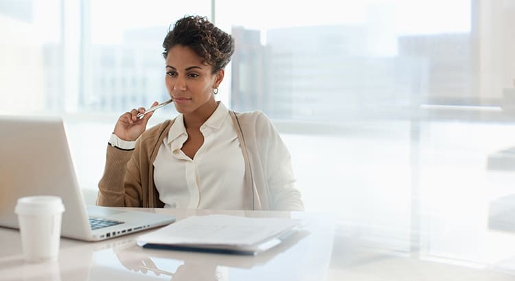 A professional woman seated at a desk, working on a laptop. She appears to be in deep thought, holding a pen to her lips as she focuses on the screen. The environment is bright and modern, with large windows allowing in natural light.