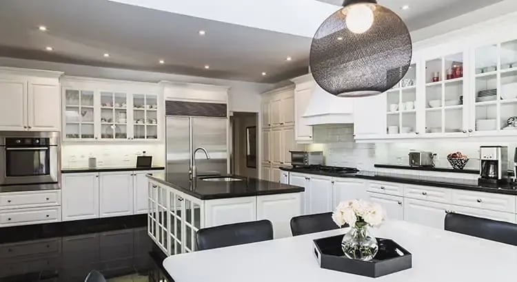 A modern, white kitchen with black countertops. The kitchen is well-lit, with a large island in the center and pendant lights hanging above.