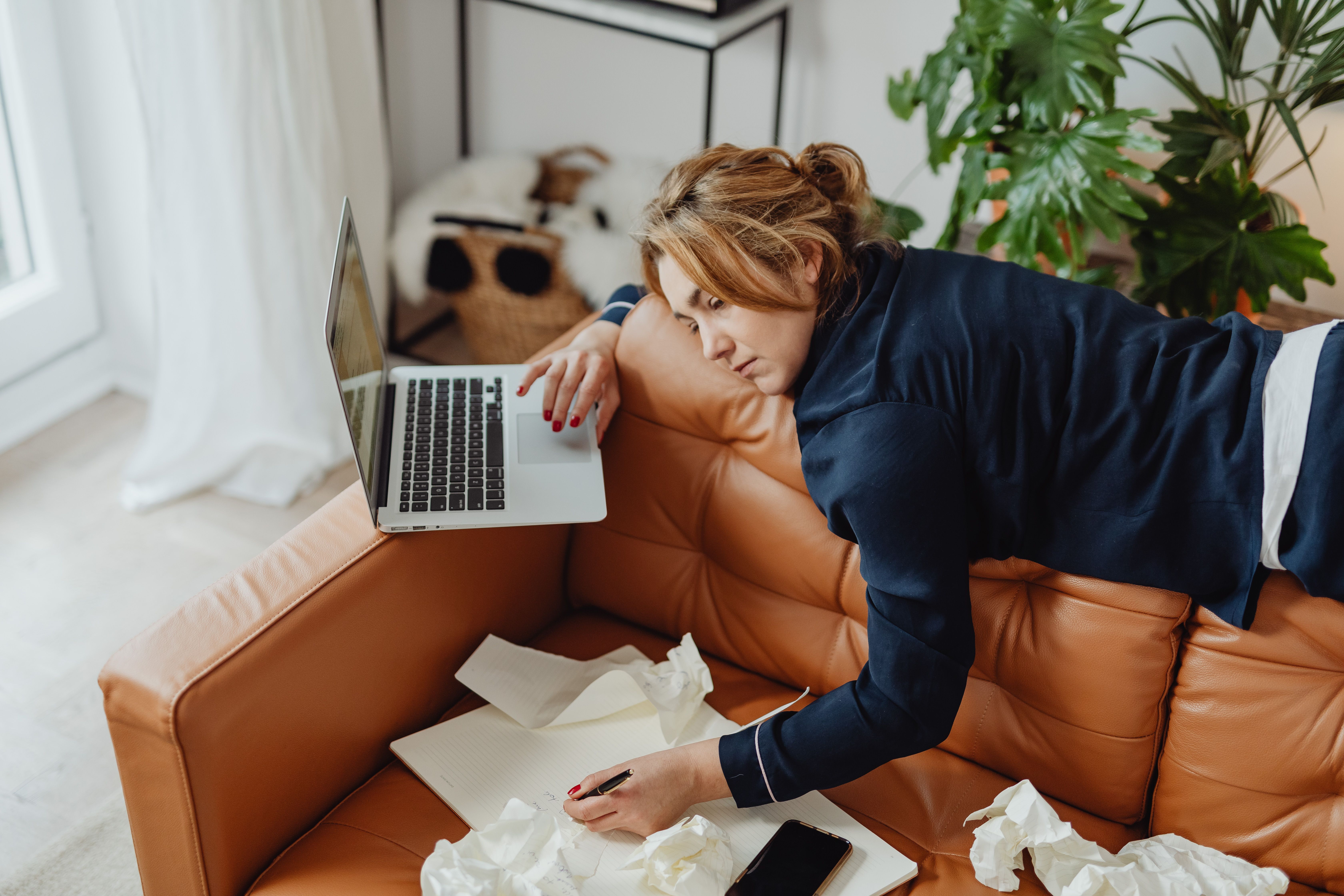 Woman writes in a notebook on a tan leather sofa beside a laptop and crumpled tissues.