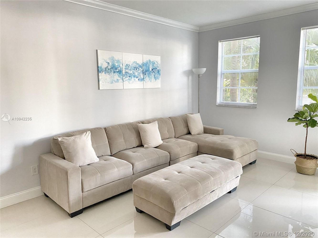 Living room with a beige sectional and tufted ottoman, blue abstract art, and bright windows.