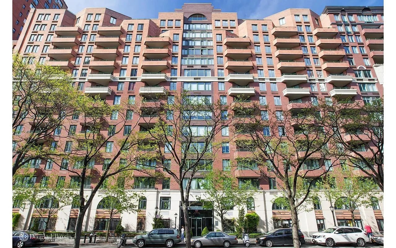 Red-brick residential building with rows of balconies, trees along the sidewalk, and parked cars.