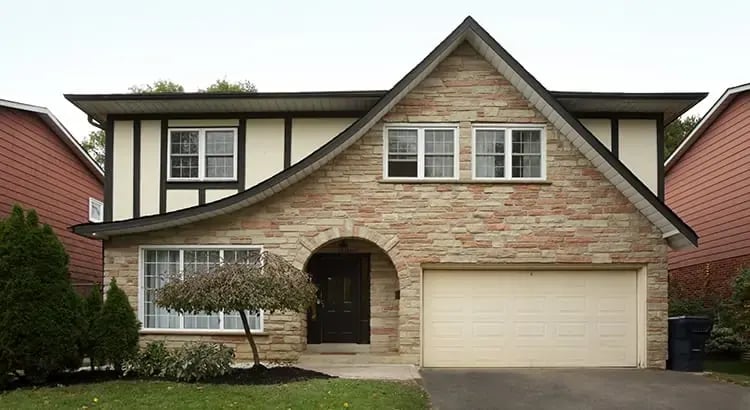 A large suburban house with a two-car garage, a well-maintained lawn, and a tree in front. The house has a gabled roof and stone exterior.