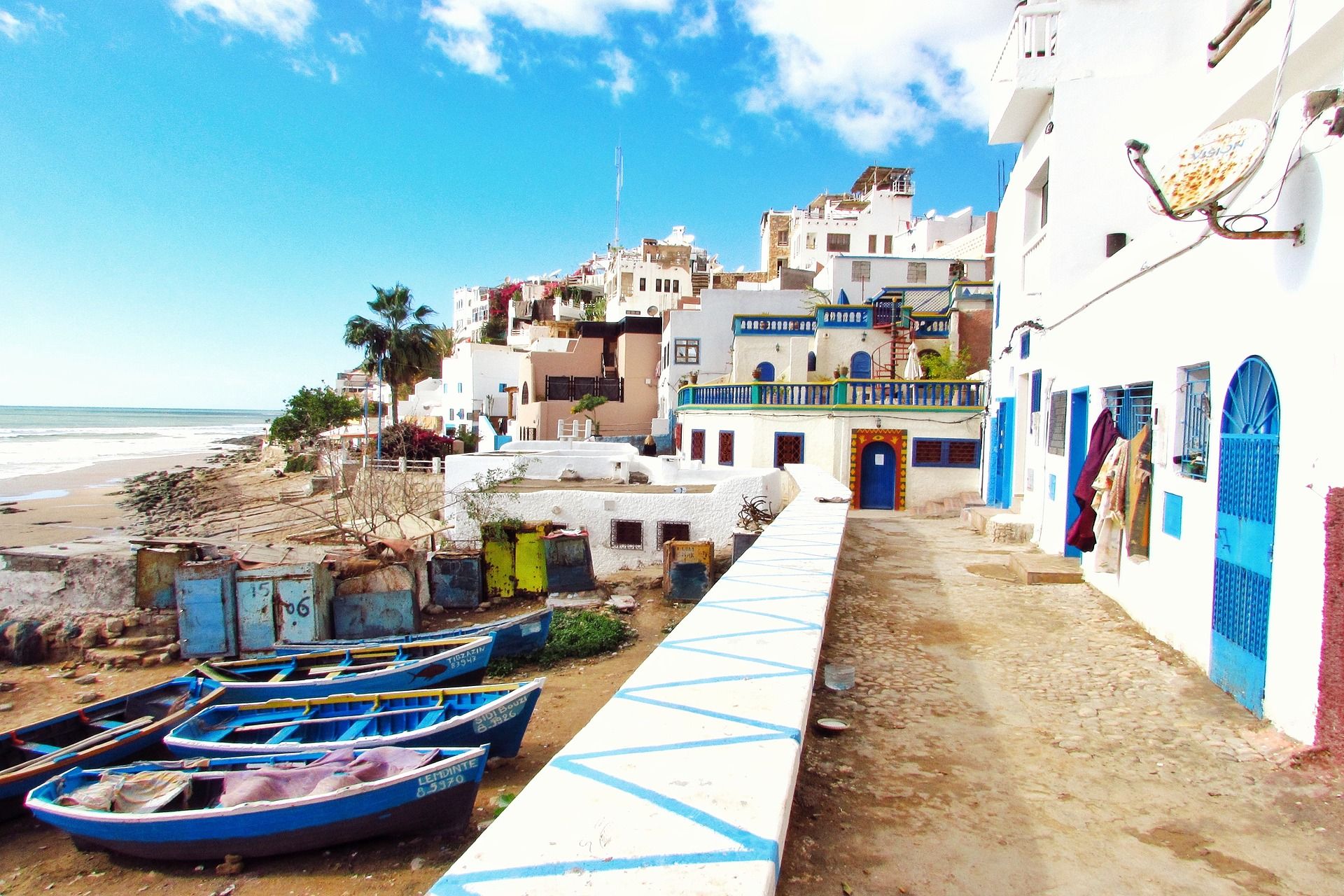 Coastal village with white buildings and blue doors, fishing boats beside a beachside walkway.