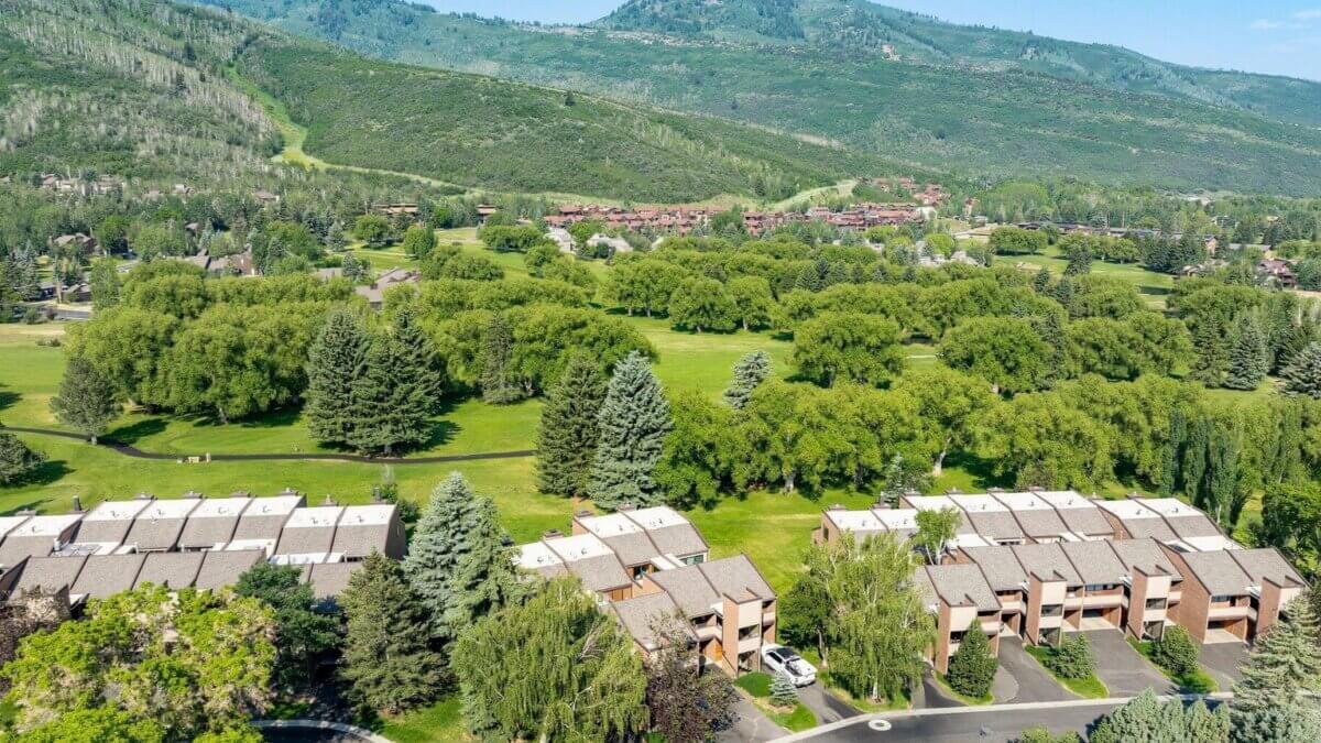 Aerial view of condominiums beside a tree-lined green park, with mountains beyond.
