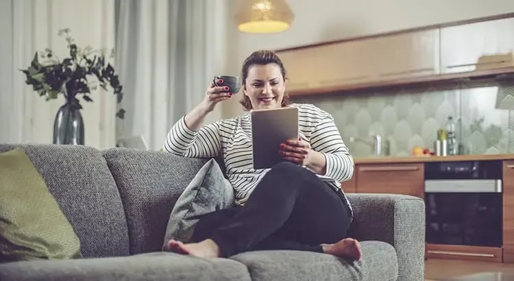 A person sitting comfortably on a couch with a tablet, drinking from a mug. The setting is a cozy living room, suggesting relaxation and leisure time.