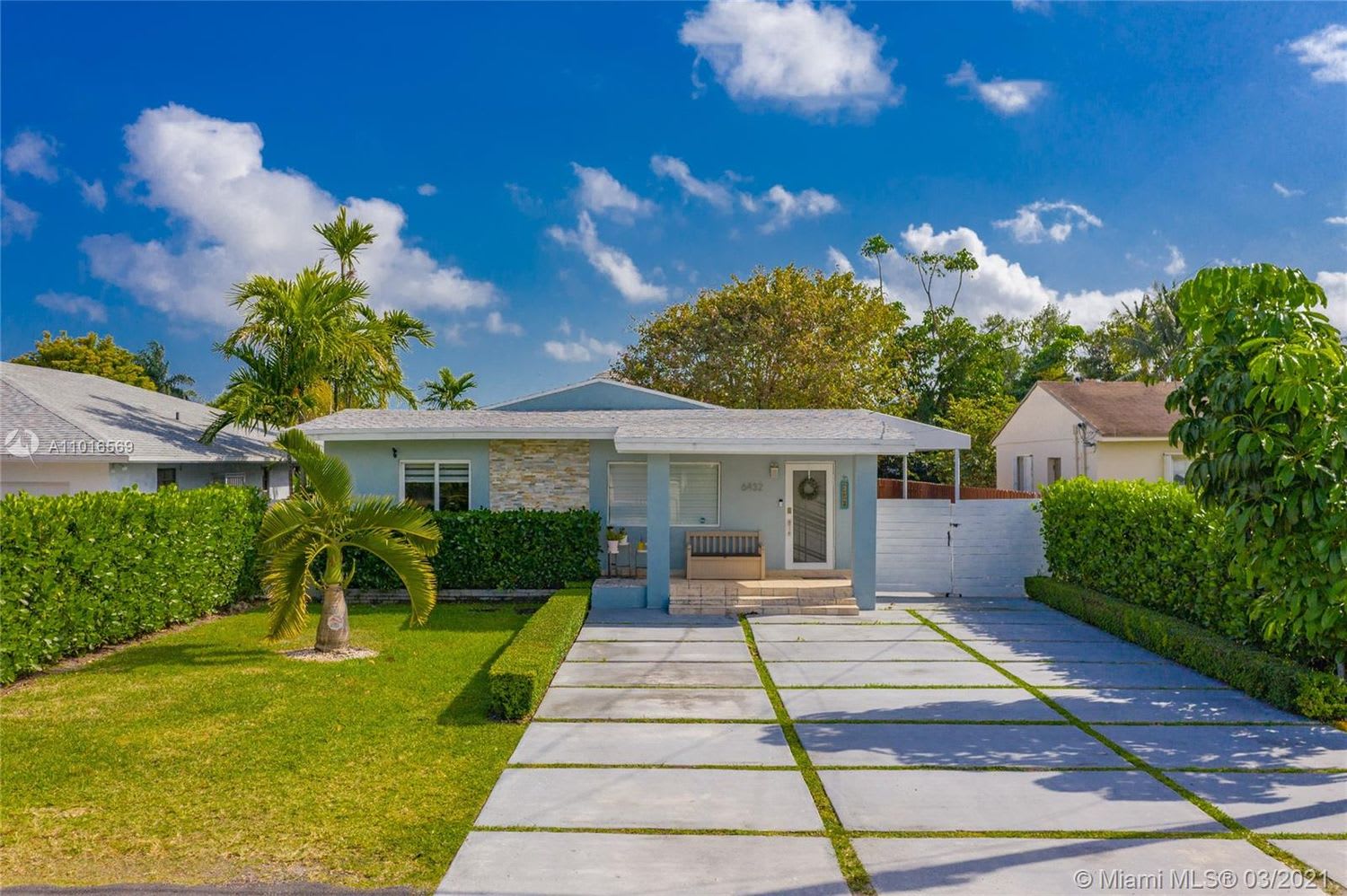 Single-story blue house with stone accents, a wide driveway, trimmed hedges, and palm trees.