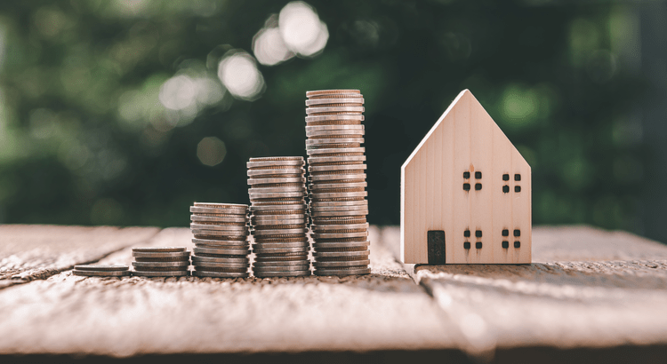 A close-up shot of stacked coins next to a small wooden house model. The background is blurred, emphasizing the foreground elements. 