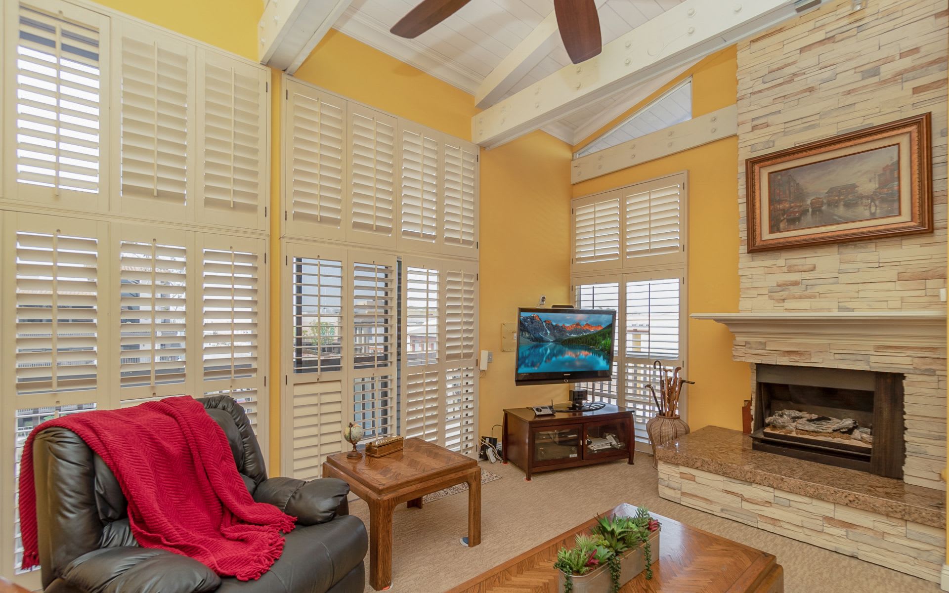 Living room with tall plantation shutters, vaulted beamed ceiling, and stone fireplace.