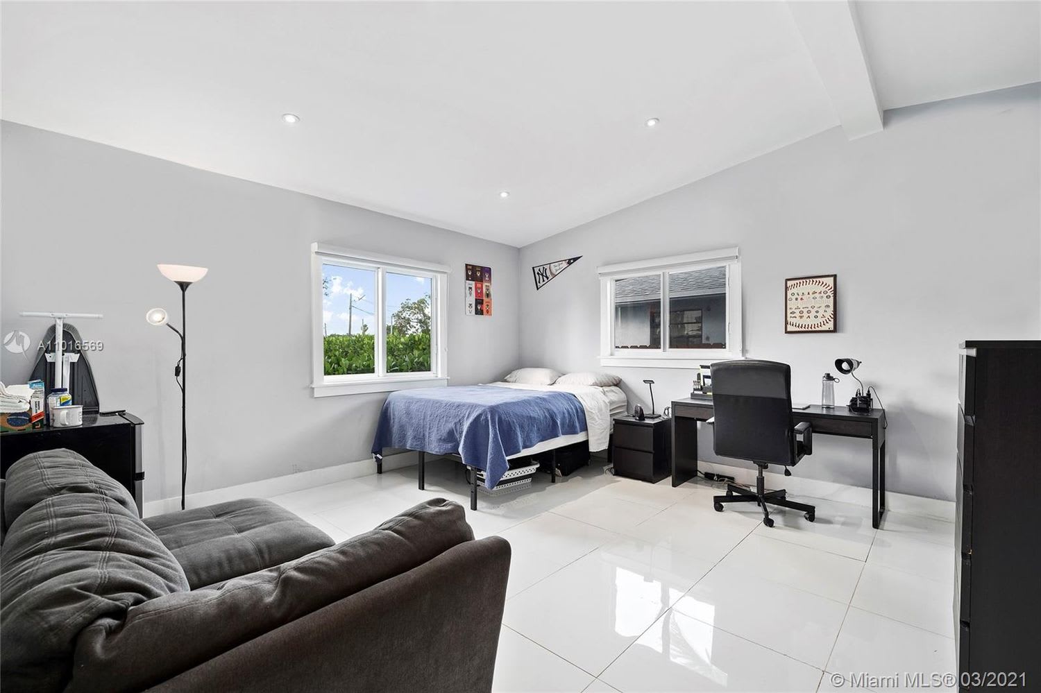 Bedroom and home office with a blue-covered bed, dark sofa, and desk beneath two windows.
