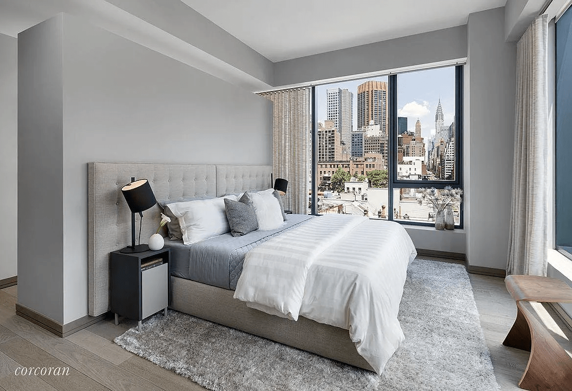 Staged bedroom with a gray upholstered bed and Manhattan skyline view through large windows.