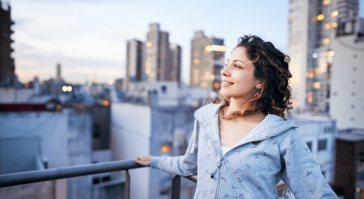 A woman standing on a balcony, looking out over a cityscape. The background shows high-rise buildings and an urban environment.