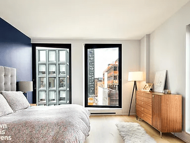 Bedroom with tall black-framed windows, a wooden dresser, and nearby city buildings outside.