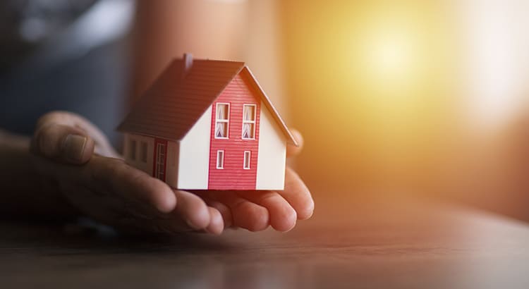 The house model is red with white details, resembling a traditional home. The background is softly blurred with a warm light source, giving the image a calm and comforting ambiance. 