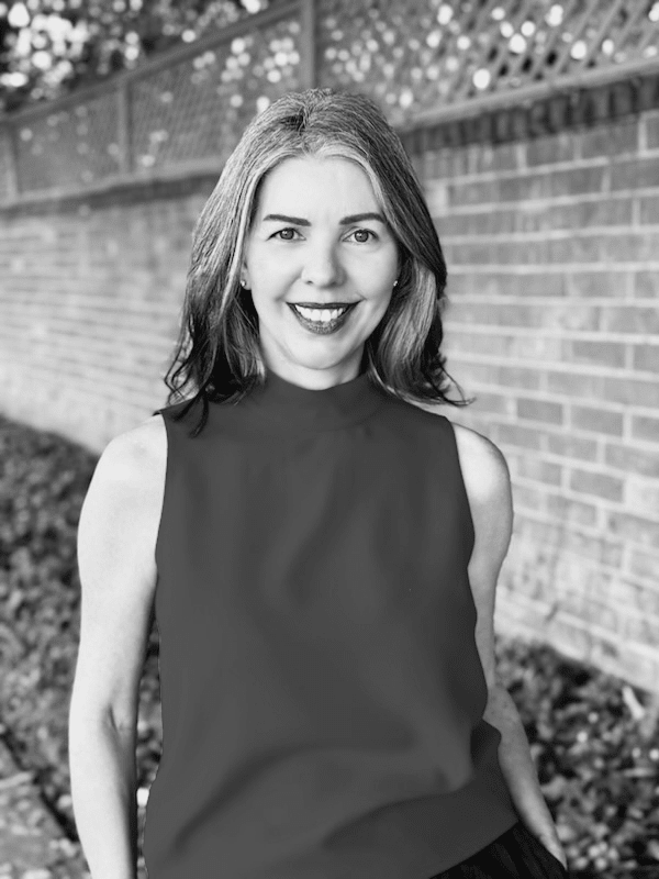 Smiling woman with shoulder-length hair in a sleeveless top, standing outdoors by a brick wall in black and white.