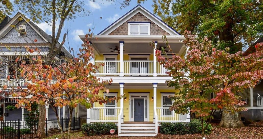 Yellow house with a sleek black metal roof, bay windows, a wooden balcony, and a crisp white picket fence.