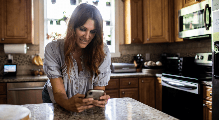 A woman stands in a kitchen, intently gazing at her phone while surrounded by cooking utensils and appliances.