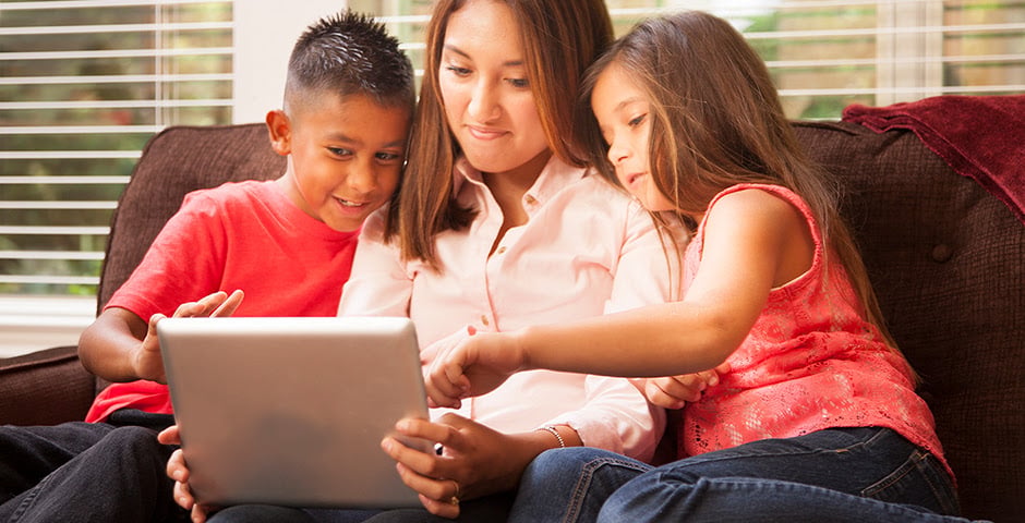 Mother and two children explore a tablet together on a living room sofa.