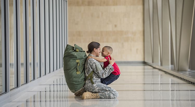 A heartwarming scene of a soldier, dressed in military uniform, embracing a young child in what appears to be an airport or similar setting. The soldier is kneeling on the ground, facing the child who is wearing a red shirt. 