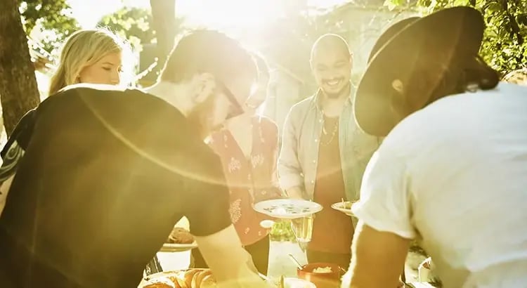 A group of people gathered outdoors, possibly at a social event or barbecue. The sunlight creates a warm and inviting atmosphere.