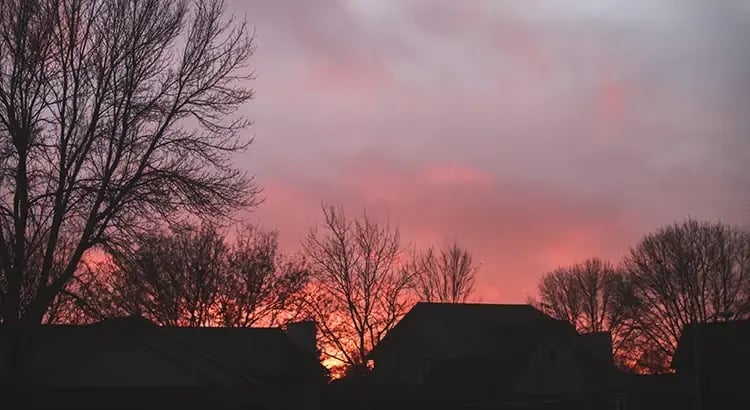  A scenic view of a sunset or sunrise with silhouettes of trees and a house in the foreground. The sky is filled with vibrant colors, creating a peaceful atmosphere.