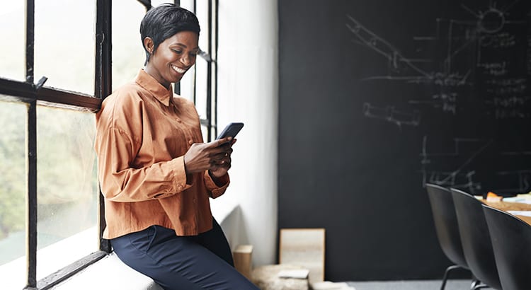 A woman sitting by a window, using her phone. She is dressed in a casual outfit and appears to be in a comfortable environment.