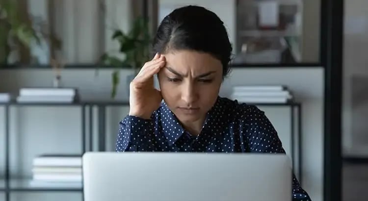 A woman sitting at a desk, looking stressed while working on her laptop. The scene suggests she is dealing with a challenging task or problem.