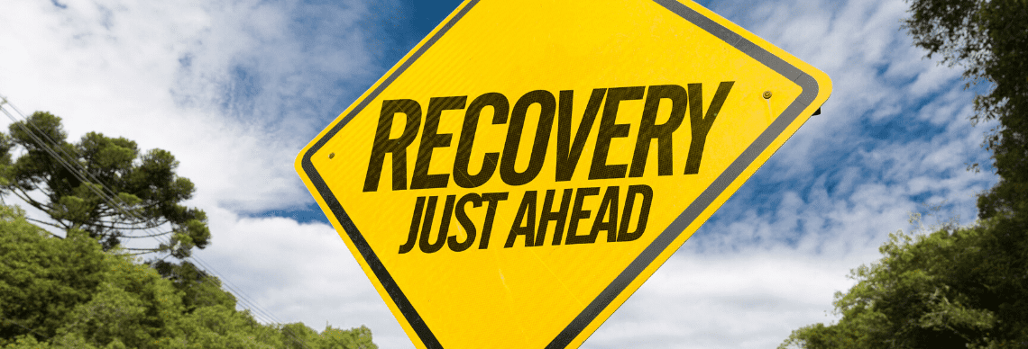 A bright yellow road sign that reads "RECOVERY JUST AHEAD" in bold black letters. The sign is set against a backdrop of a clear sky with scattered clouds and lush green trees on either side, suggesting a natural or outdoor setting.