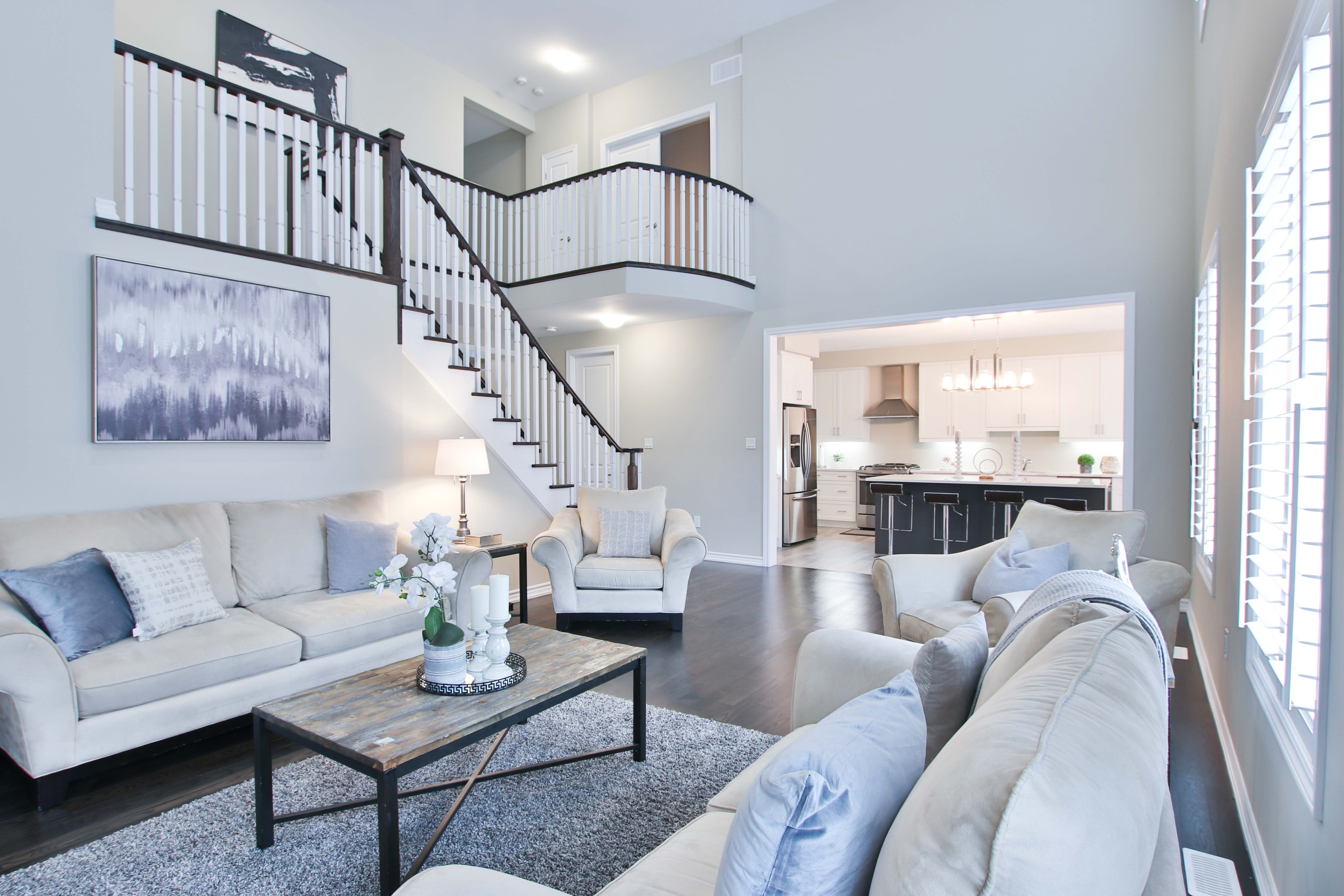 Staged living room with pale sofas, a dark-railed staircase, and an open kitchen.