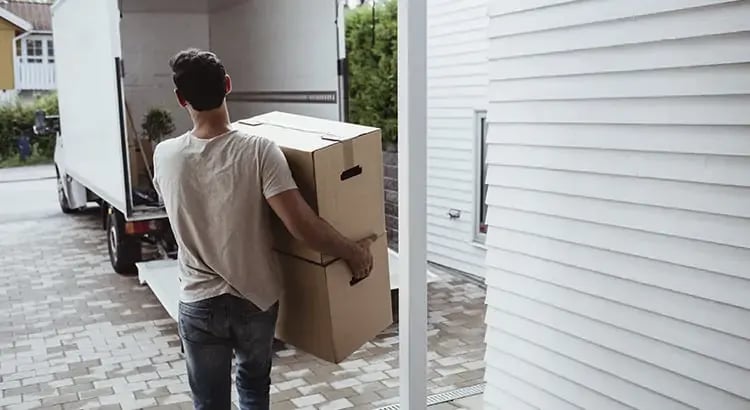 A person carrying a large cardboard box, possibly moving into or out of a house. The scene includes a white house and a driveway, indicating a moving or relocation process.