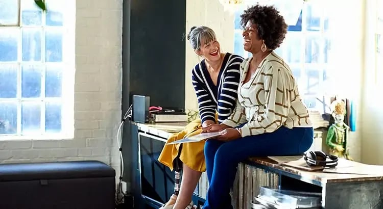 Two women sitting on a countertop in a bright, casual setting. Behind them is a large, multi-pane window letting in natural light, illuminating the room.