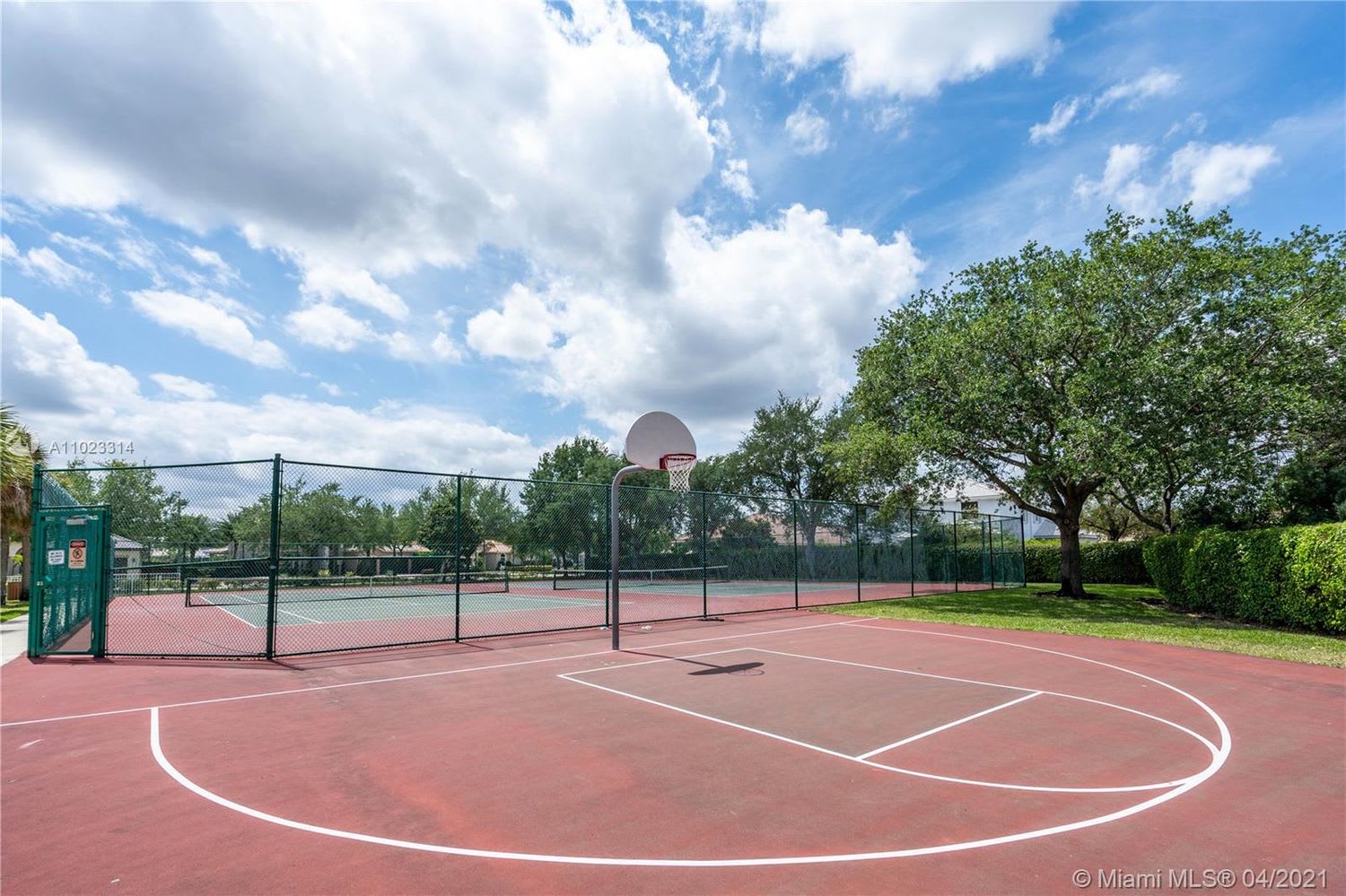 Outdoor basketball court with red paving, chain-link fencing, and mature trees.
