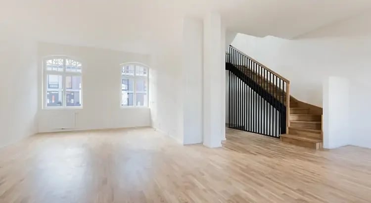 An empty, well-lit room with light wood flooring. There is a staircase with dark railings leading to an upper floor, and large windows letting in natural light.