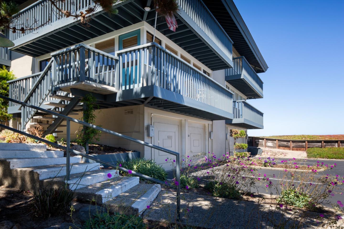 Multi-level home with blue-gray balconies, broad concrete steps, and purple flowers along the entry path.