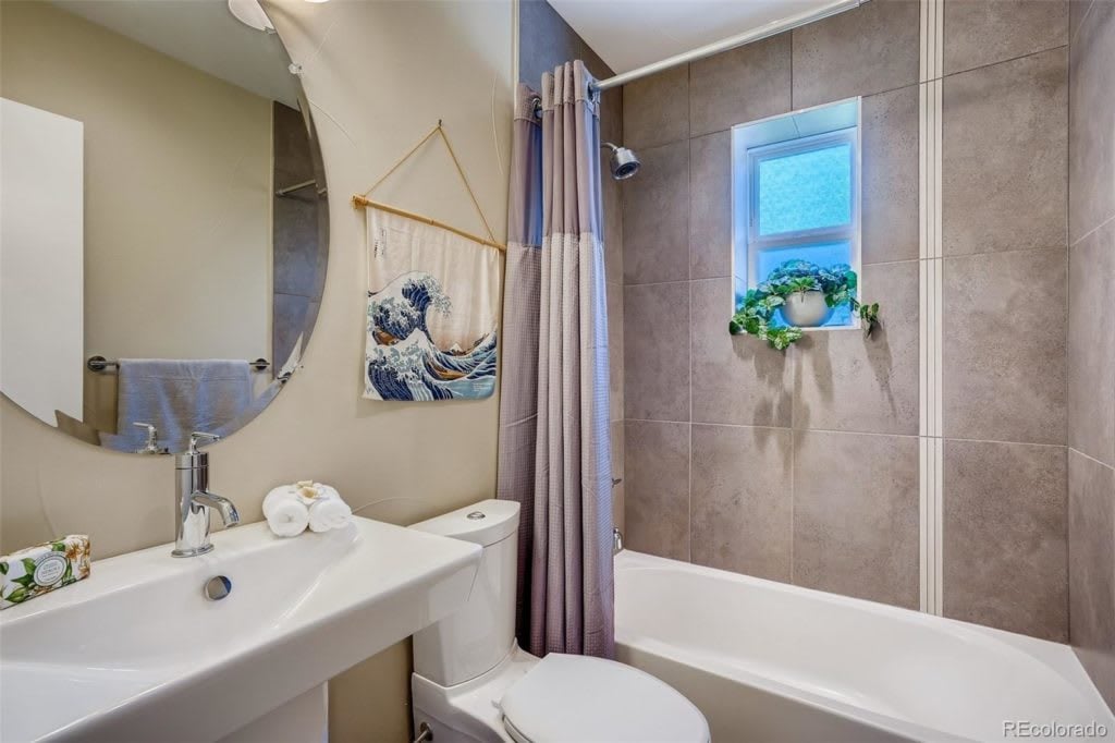Bathroom with tiled tub and shower, pedestal sink, round mirror, and a small window with plants.