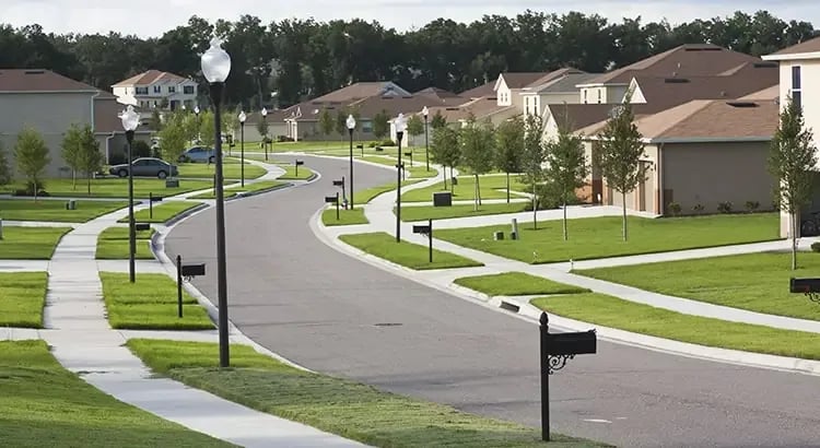 A suburban neighborhood street with neatly maintained lawns, streetlights, and houses lined up along the road. The image showcases a peaceful residential area.