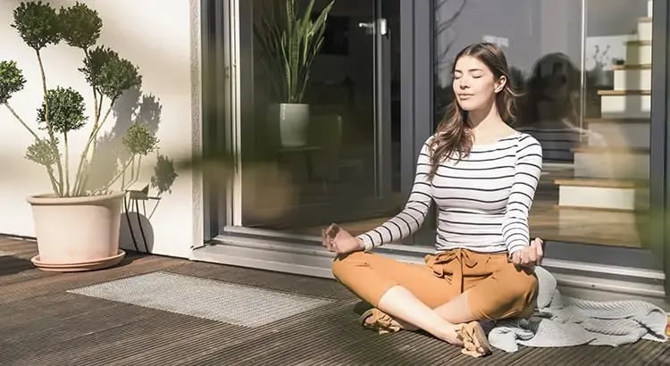 A woman sitting cross-legged in a meditative pose on a porch or balcony. She is surrounded by plants and appears to be practicing mindfulness or relaxation.